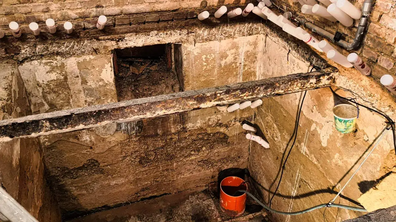 Arbeiten am **Naturkeller Boden sanieren**: Feuchte Wände, Rohre und ein Eimer deuten auf eine Baustelle hin.