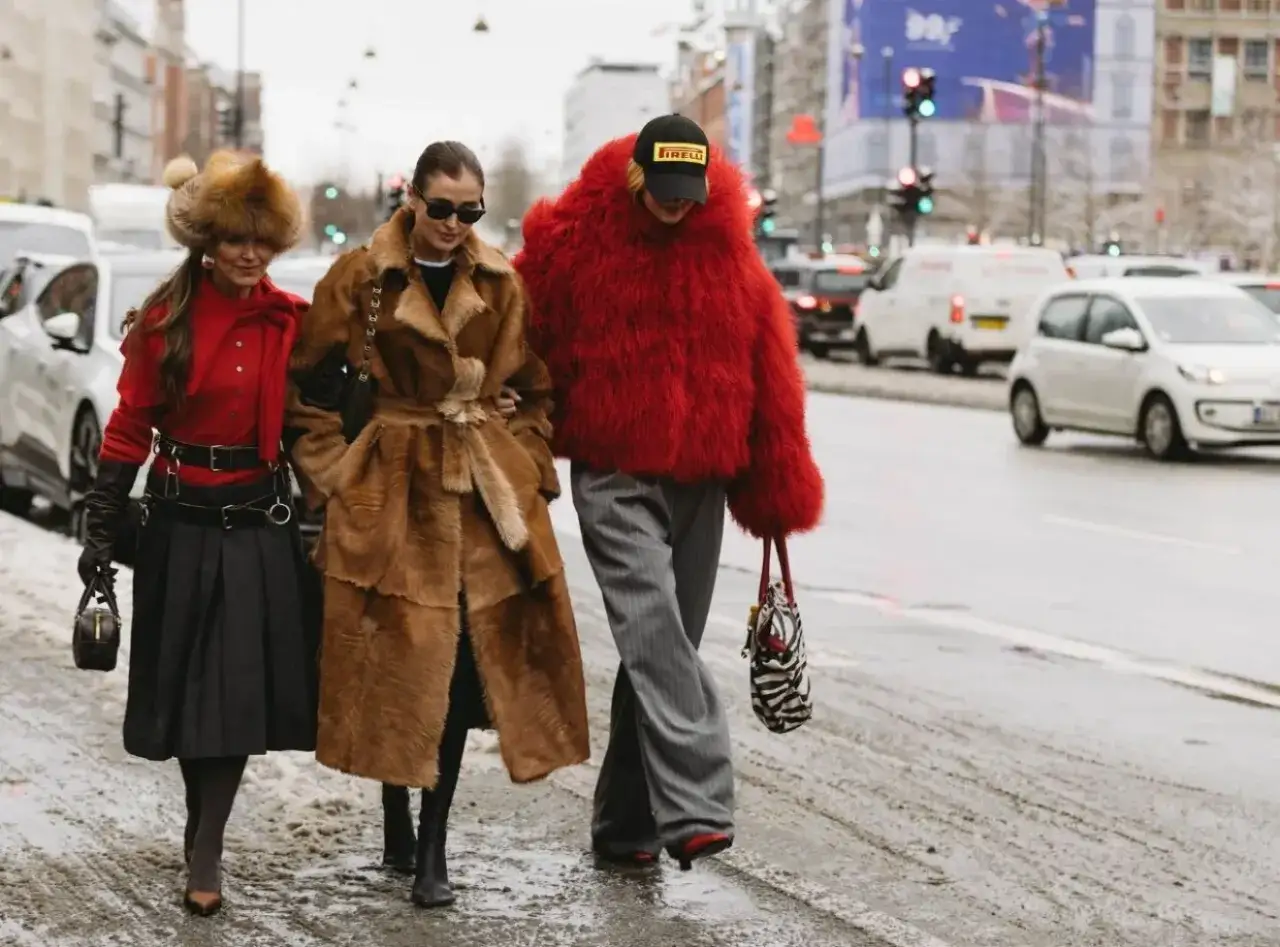 Tre kvinnor i trendiga vinterkläder, en med röd pälsjacka, visar upp sin copenhagen street style på en snöig gata.