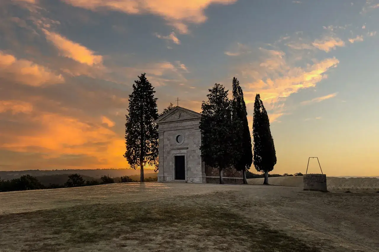 Piccola chiesa val d'Orcia al tramonto, con cipressi e un pozzo antico. Cielo infuocato.