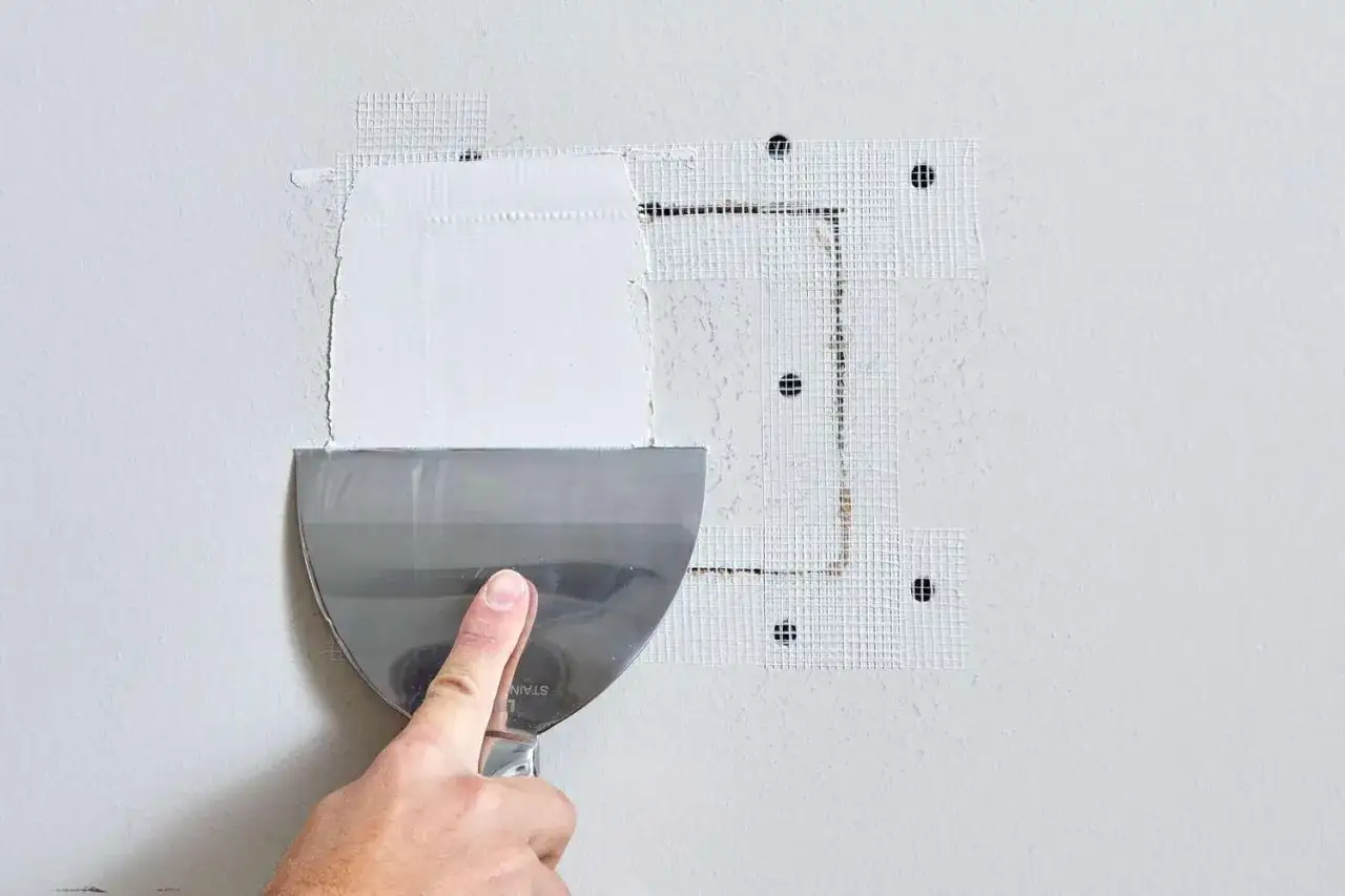 Aplicando masilla con esp&aacute;tula para tapar agujeros en la pared, usando malla de fibra de vidrio para reforzar.