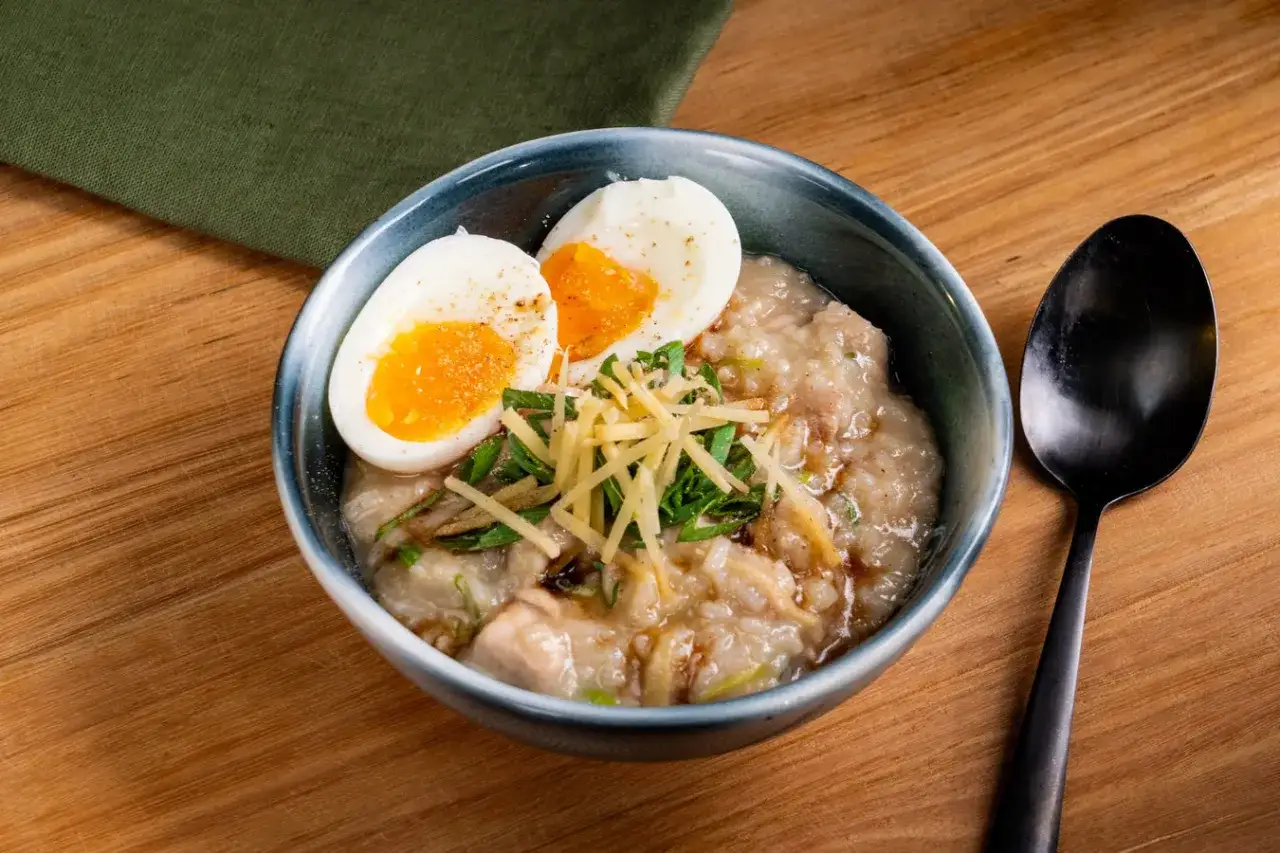Herzhaftes Congee Rezept mit weichgekochtem Ei, Ingwer und Fr&uuml;hlingszwiebeln in einer Sch&uuml;ssel auf Holztisch.