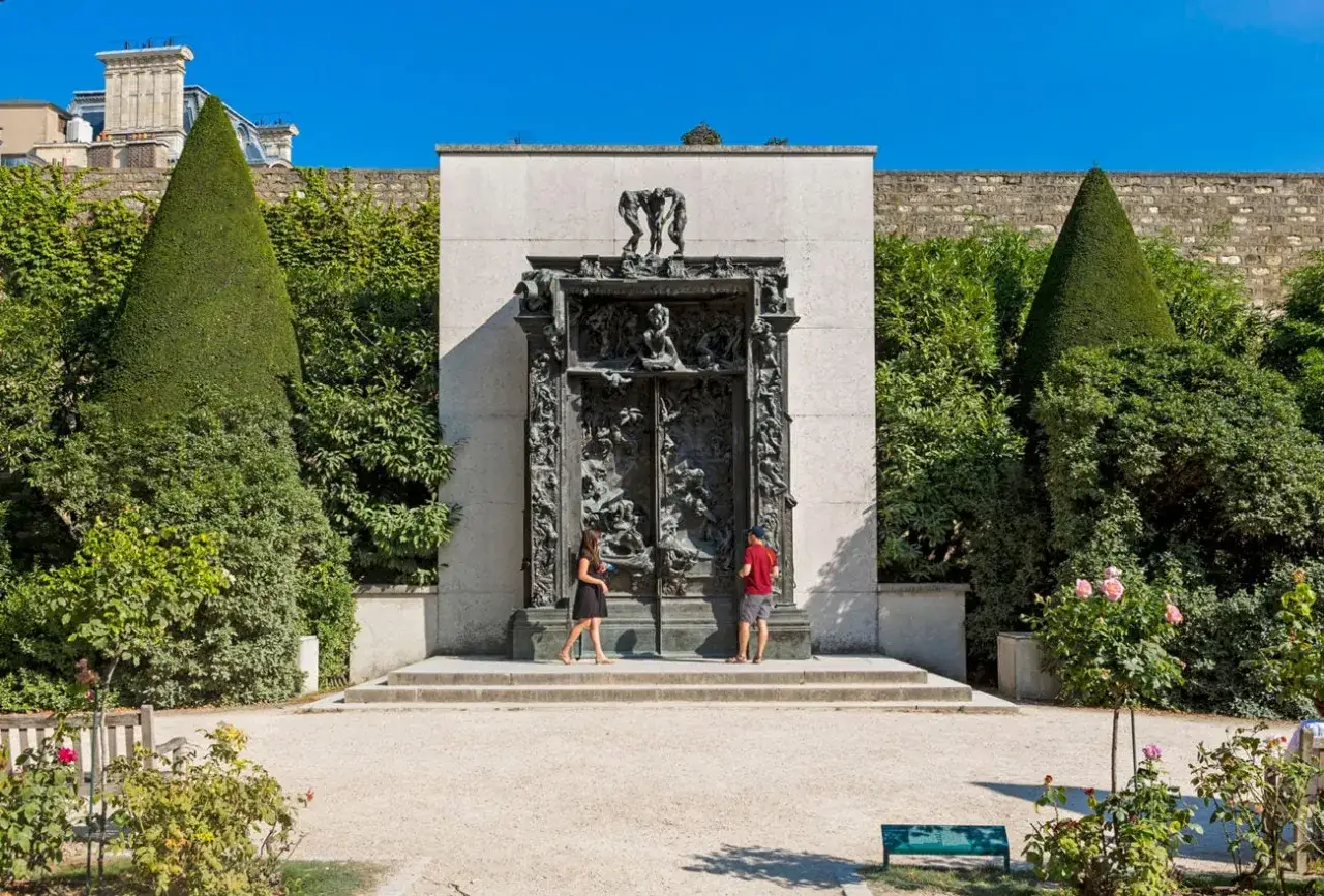 Deux visiteurs contemplent la Porte de l'Enfer, une œuvre monumentale de Rodin, dans un jardin verdoyant sous un ciel bleu éclatant.