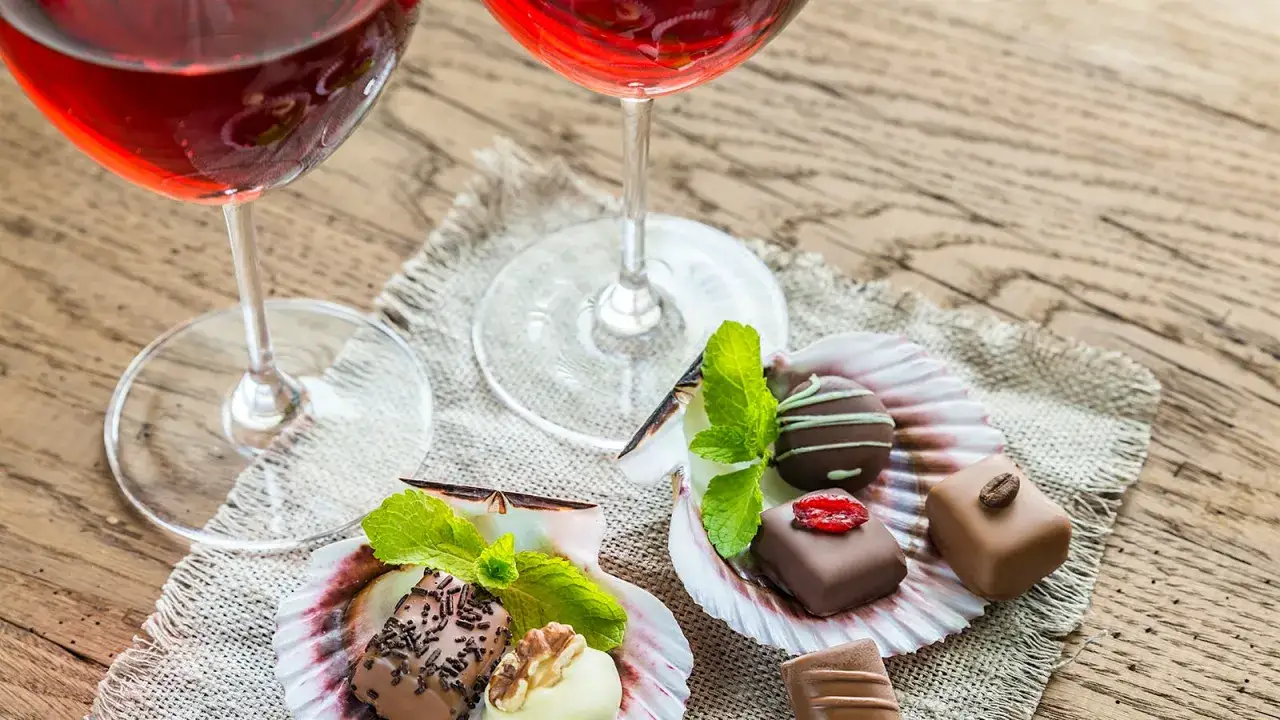 Zwei Gl&auml;ser Rotwein stehen neben einer Auswahl an exquisiten Pralinen und Schokoladen, garniert mit Minze und N&uuml;ssen.