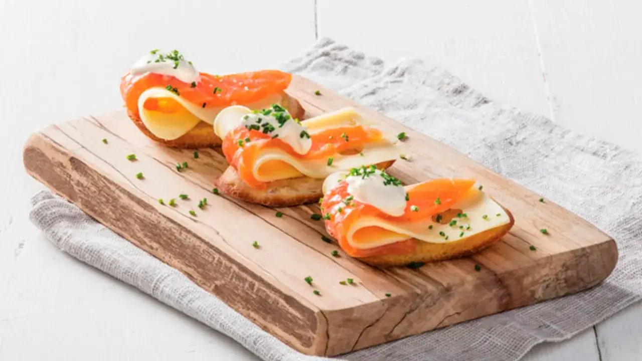 Tres canap&eacute;s de salm&oacute;n ahumado, queso y crema sobre pan tostado, adornados con cebollino, sobre una tabla de madera.