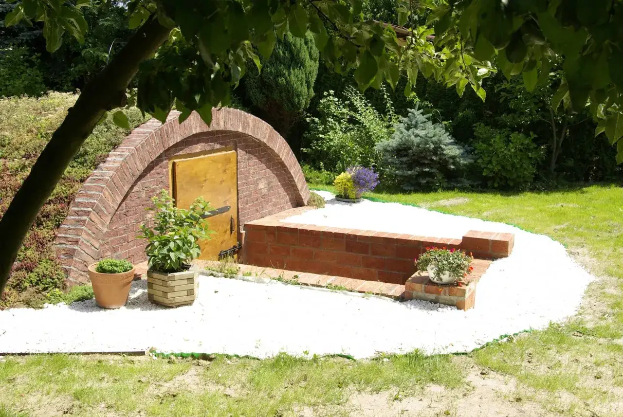 Piwnica ogrodowa z cegły, z drewnianymi drzwiami, otoczona białym żwirem i zielenią. Zastanawiasz się, ile kosztuje piwnica ogrodowa?