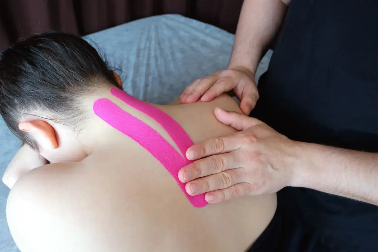 Różowe tejpy na szyję i bark pomagają w rehabilitacji. Terapeuta delikatnie uciska skórę.
