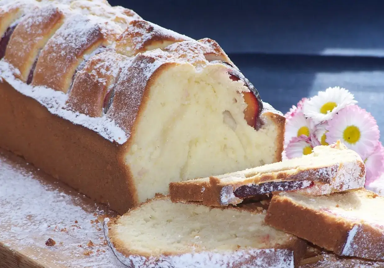 Szybkie ciasto z 4 składnik&oacute;w, pyszny keks z kawałkami śliwek, posypany cukrem pudrem, obok bukiecik stokrotek.