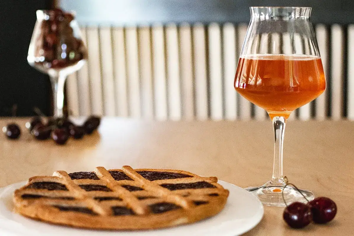 Un bicchiere di birra alla ciliegia ambrata accanto a una crostata e ciliegie fresche.