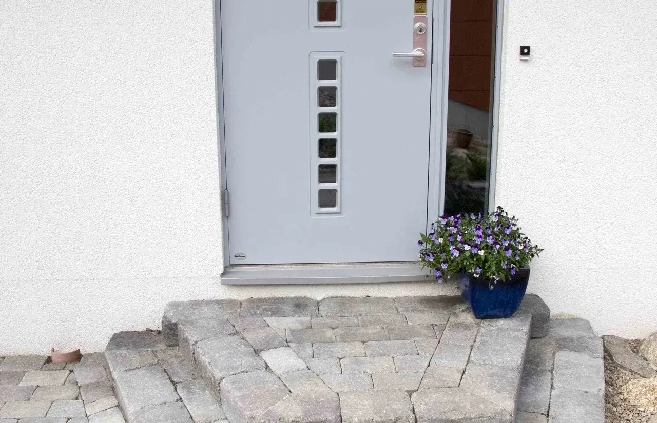 En vacker stentrappa leder upp till en modern ytterd&ouml;rr. En bl&aring; kruka med lila blommor st&aring;r vid sidan.