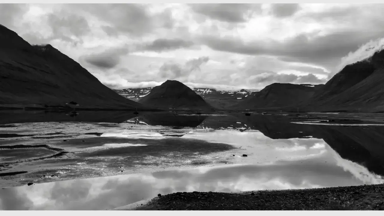 A vast, reflective pan f of a fjord with dramatic mountains and a cloudy sky.