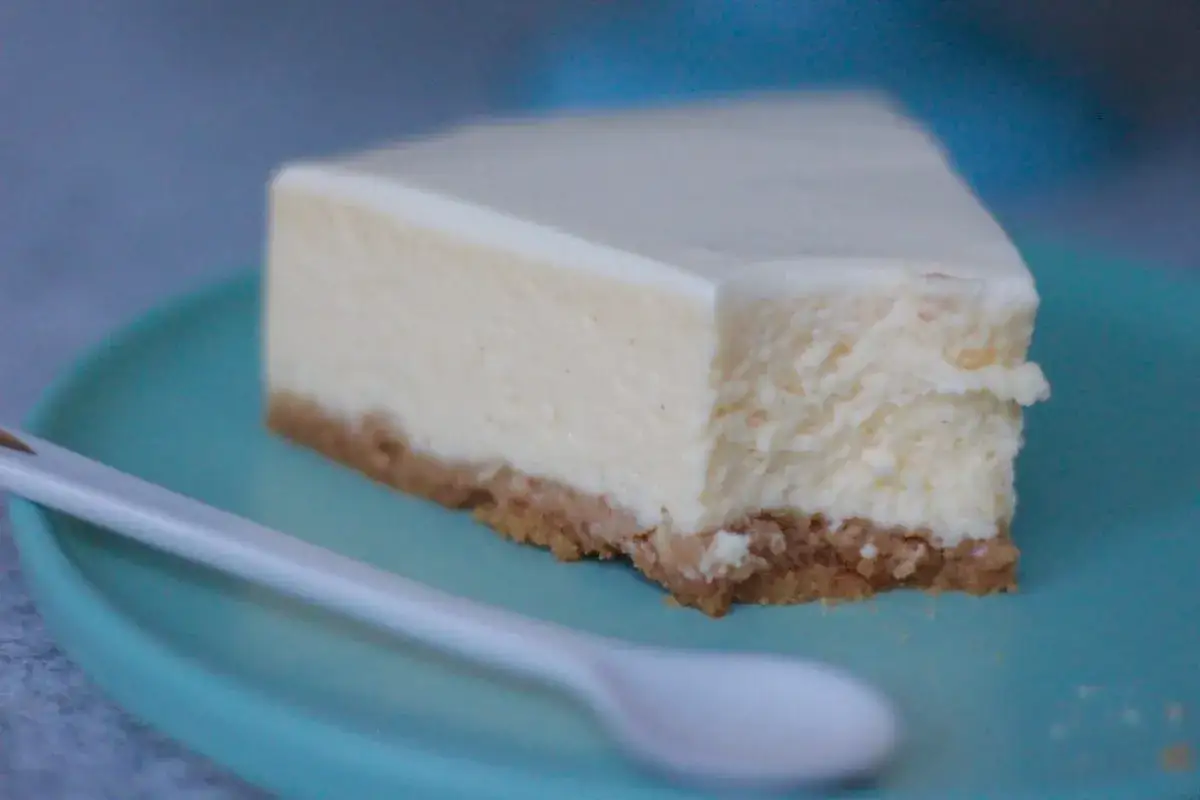 Ein Stück New York Cheesecake mit Keksboden auf einem blauen Teller, daneben ein weißer Löffel.