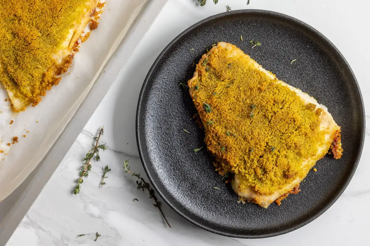 Filetto di persico dorato e croccante, appena sfornato, servito su un piatto nero con rametti di timo.
