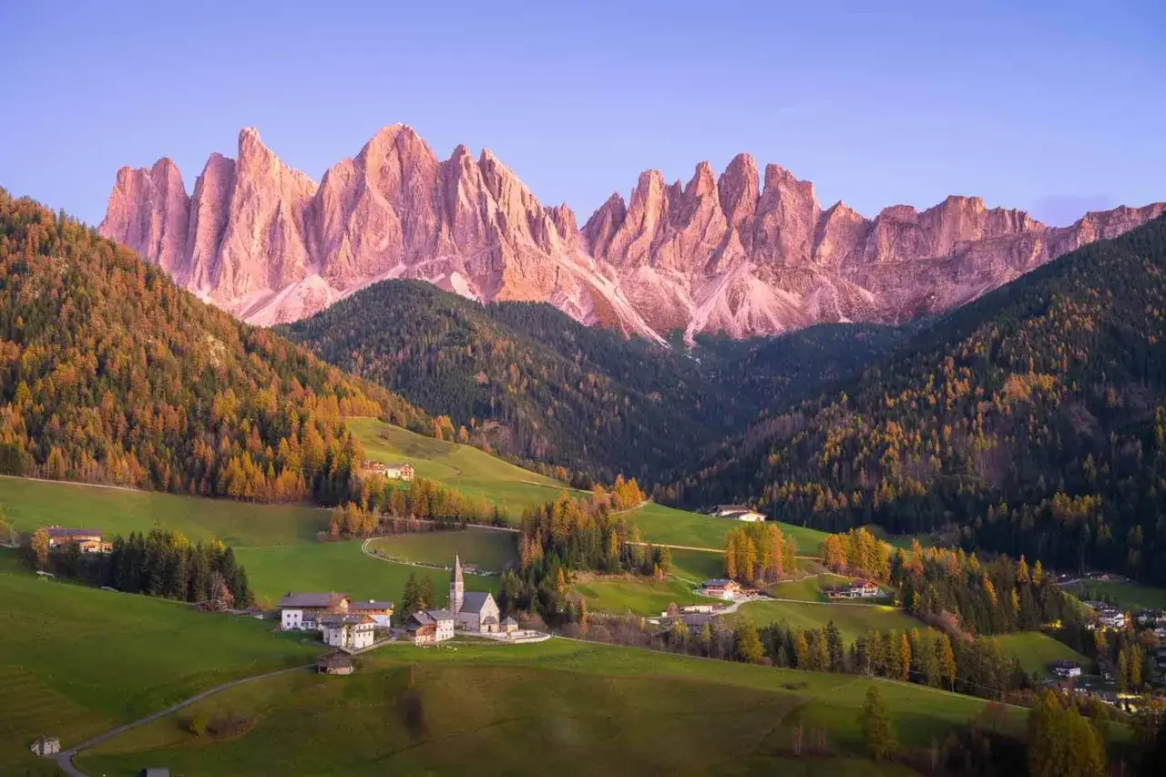Charming Italy mountain towns nestled in a valley, with a church steeple rising above the rooftops, framed by colorful autumn trees and dramatic pink-hued peaks.