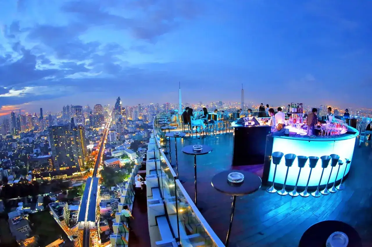 En takbar i Thonglor, Bangkok, med en fantastisk utsikt över staden i skymningen. Folk sitter vid bord och baren är upplyst i blått.