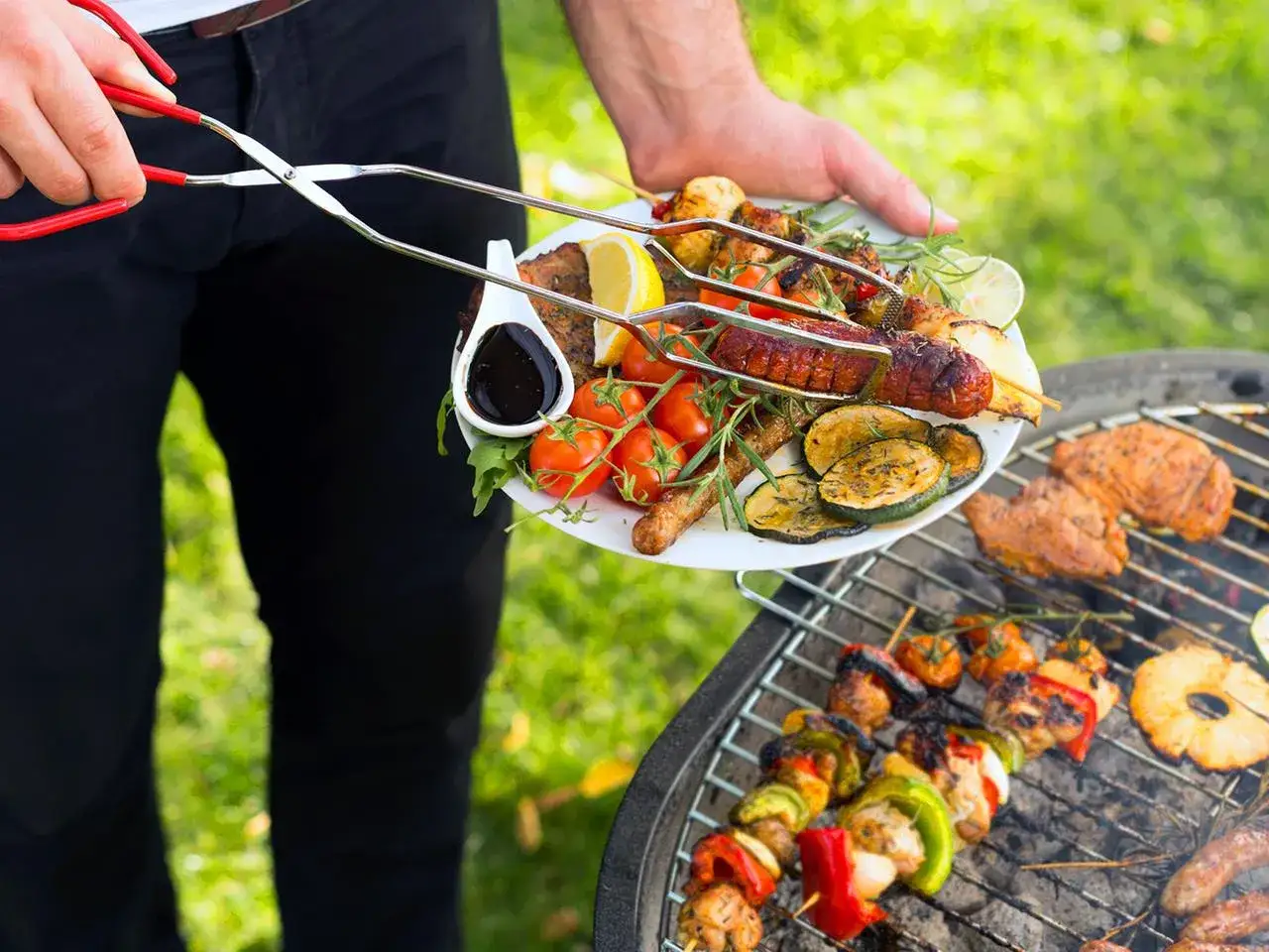 Leckere Grillideen: Ein Teller mit Würstchen, Hähnchenspießen, Gemüse und Zitronenspalten wird vom Grill genommen.