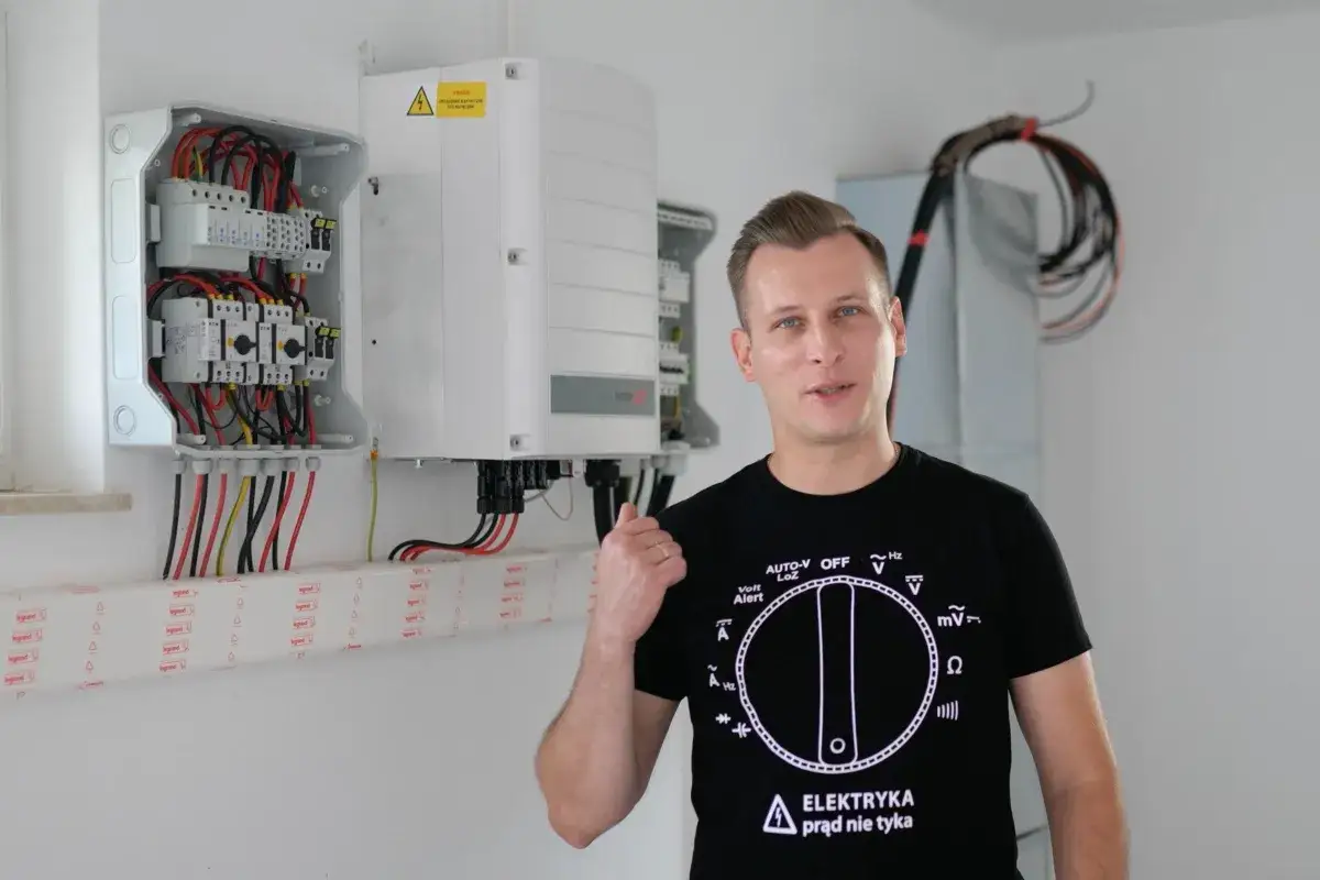 Mężczyzna w koszulce z symbolem miernika pokazuje na skrzynkę z bezpiecznikami i falownik, gdzie można regulować ampery.