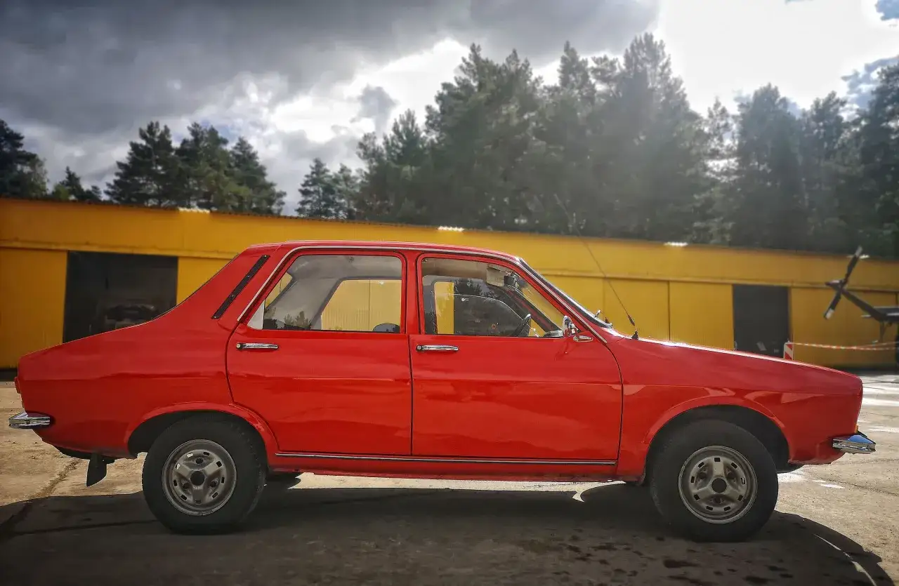 Czerwona Dacia 1300 stoi przed żółtym budynkiem, na tle drzew i burzowego nieba.