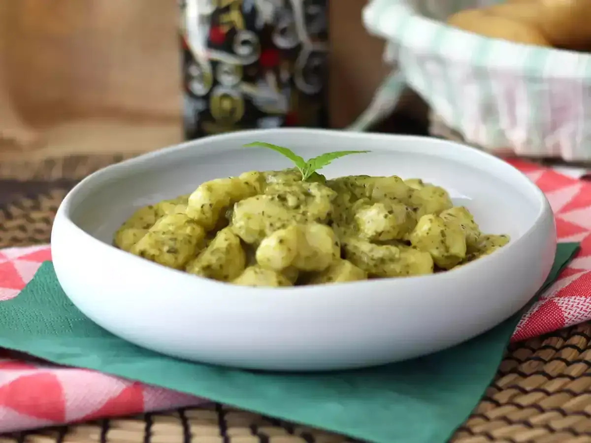 Un plat de gnocchi pesto cr&eacute;meux, garni d'une feuille de basilic, servi dans un bol blanc sur une serviette verte et un torchon &agrave; carreaux rouges.