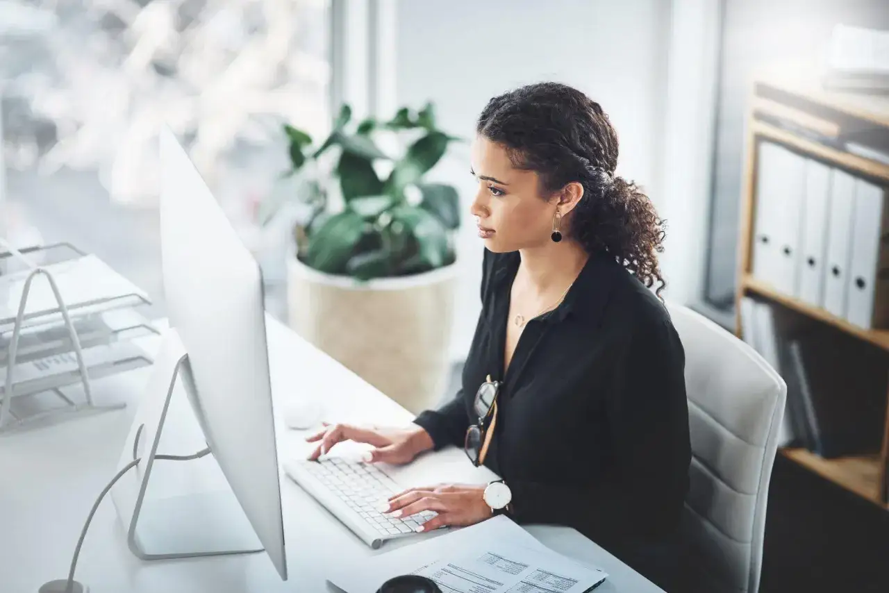 Secr&eacute;taire concentr&eacute;e sur son ordinateur, peut-&ecirc;tre en train de consulter une liste maladie professionnelle. Bureau moderne avec plante et &eacute;tag&egrave;res.