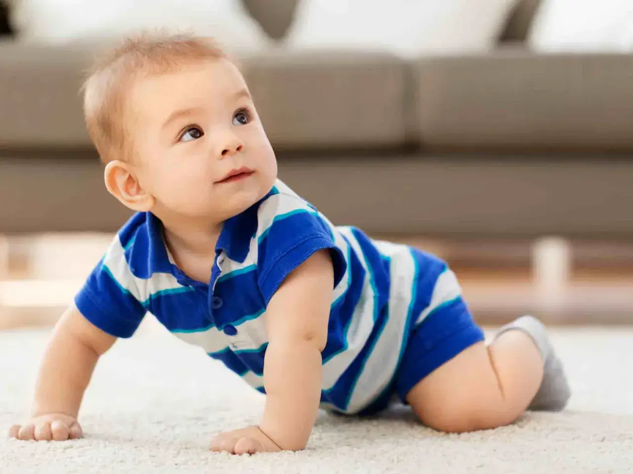 Un adorabile neonato di 8 mesi, vestito con una polo a righe blu e bianche, gattona su un tappeto chiaro, esplorando il mondo con curiosit&agrave;.