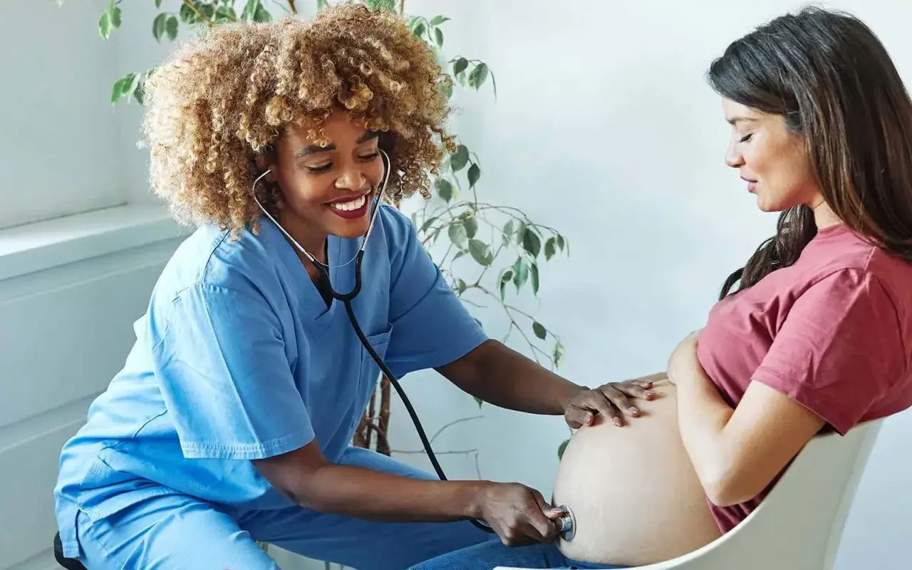 Eine Hebamme mit Stethoskop untersucht den Bauch einer schwangeren Frau. Ein sch&ouml;nes Bild, das die F&uuml;rsorge und den Wert der Hebammenarbeit zeigt, die sich auch im Hebamme Gehalt widerspiegelt.