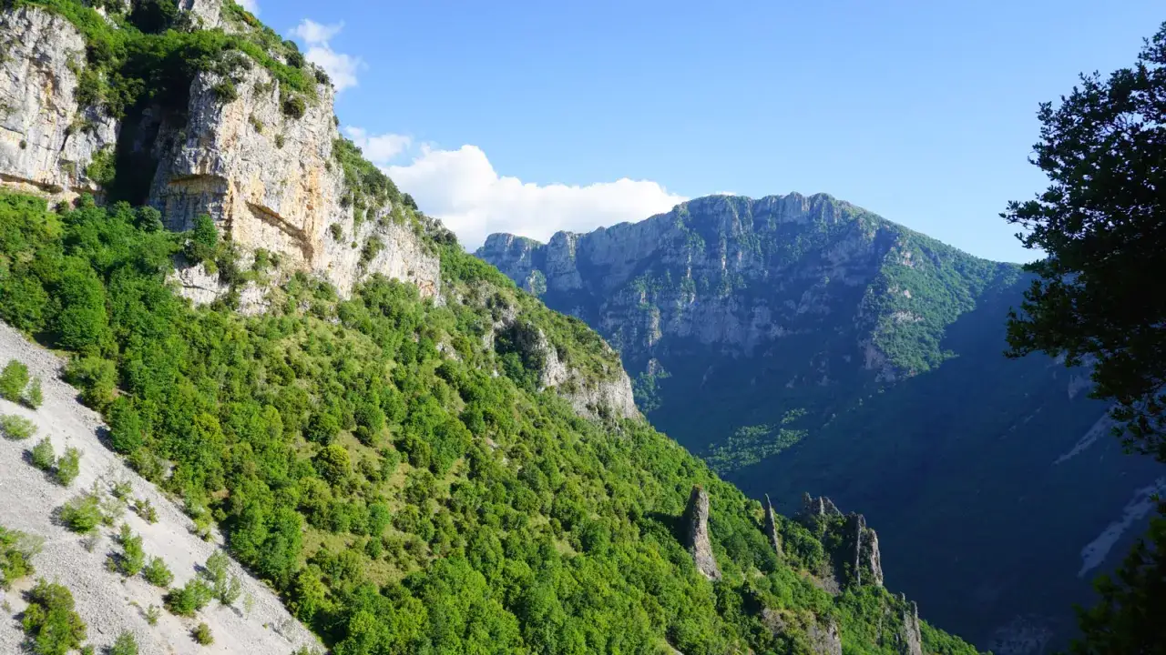 A breathtaking view of a lush green mountain valley, perfect for hiking in Greece. Jagged cliffs rise on either side, with a clear blue sky above.