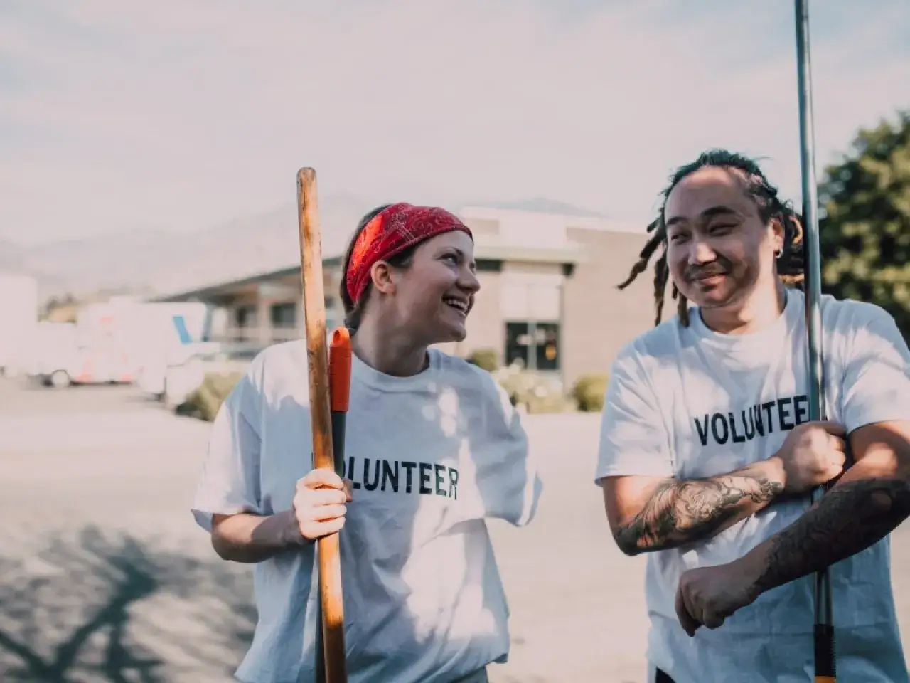 Two smiling volunteers in matching t-shirts take a break from their work, holding gardening tools.