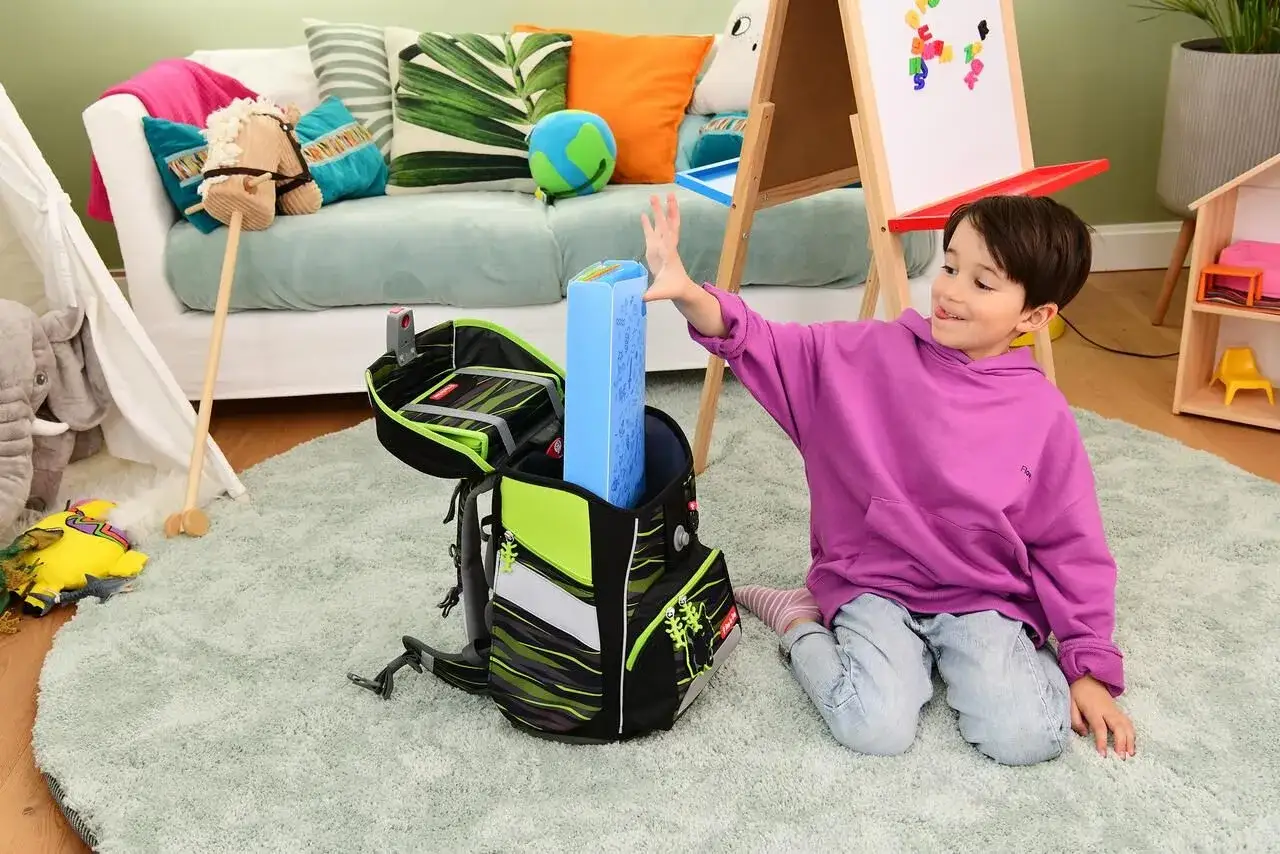 Junge packt Schulranzen für einen guten Schulstart nach den Ferien. Bunter Ranzen, Spielzeug und Tafel im Hintergrund.