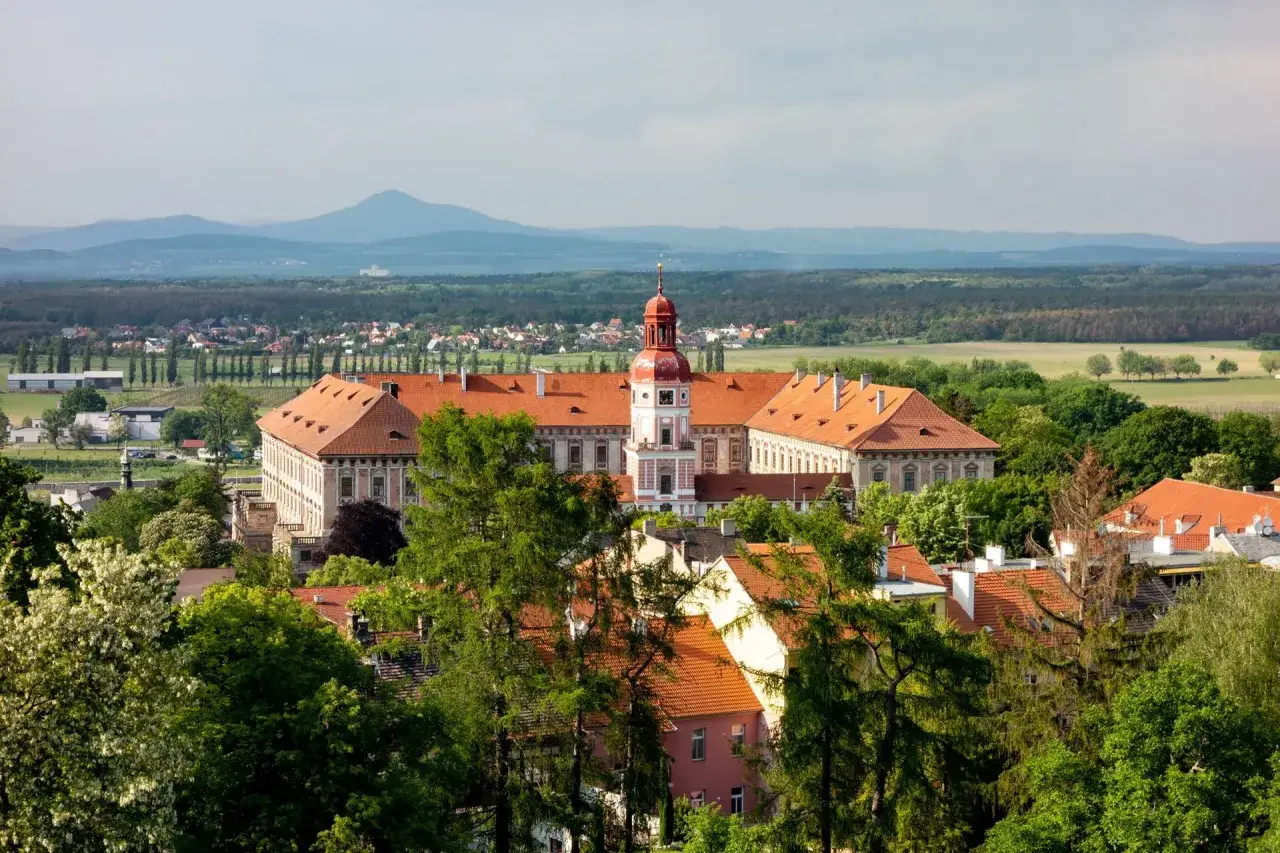 Schloss Roudnice nad Labem mit roten Ziegeld&auml;chern und einem markanten Turm, umgeben von gr&uuml;nen B&auml;umen und einer h&uuml;geligen Landschaft.