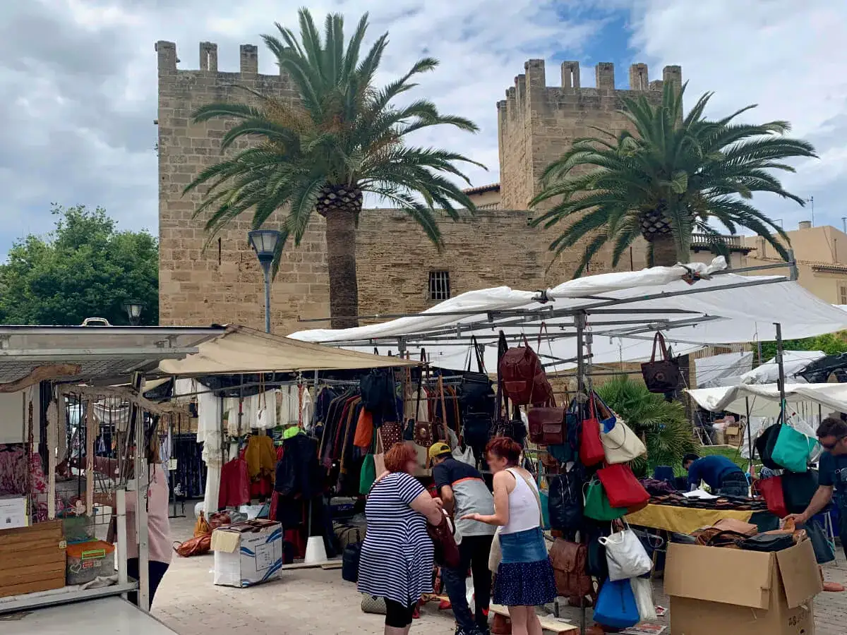 Bunte Taschen und Kleidung h&auml;ngen an St&auml;nden auf dem alcudia markt vor einer alten Burgmauer und Palmen.