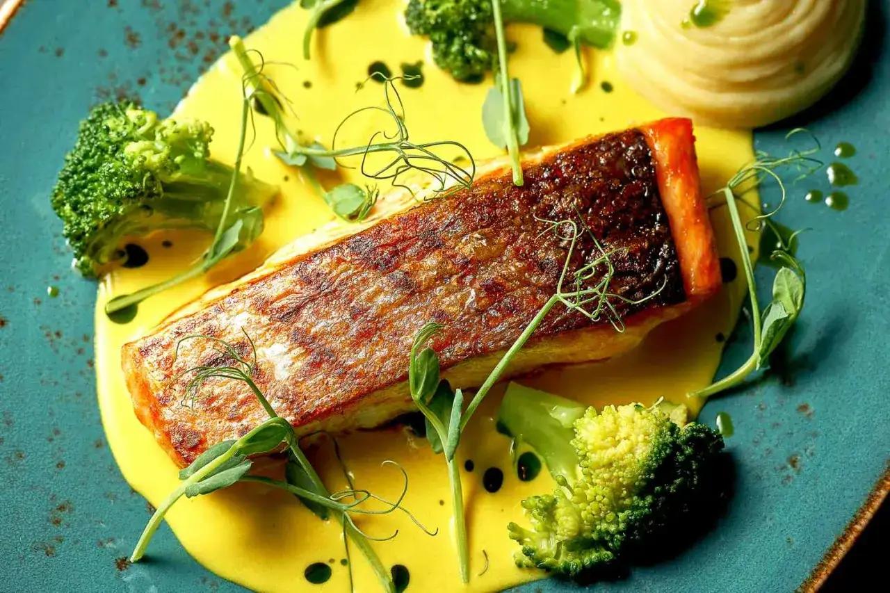 Delicioso salmón a la plancha con piel crujiente, servido sobre una cremosa salsa amarilla, acompañado de brócoli y puré.