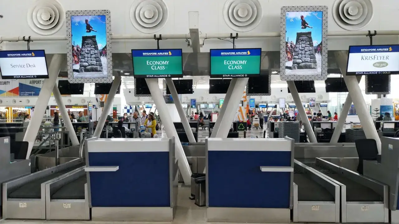 Mobywatel na lotnisku, w tle stanowiska odprawy Singapore Airlines. Na ekranach wyświetlane są informacje o klasie ekonomicznej i programie KrisFlyer.