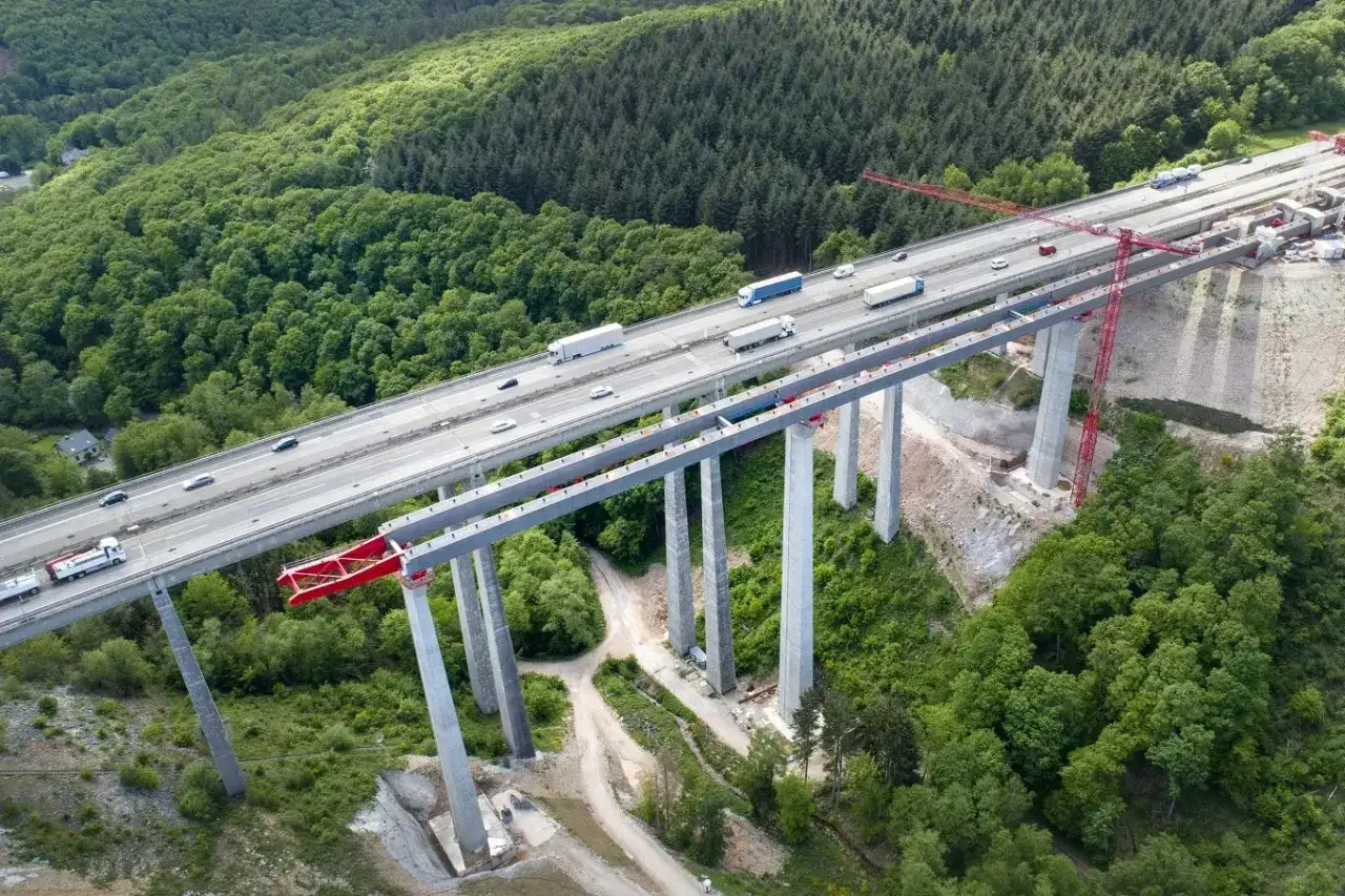 Most wysoki, z betonem C30/37, przecina zielony las. Po jego pasach poruszają się ciężarówki i samochody.