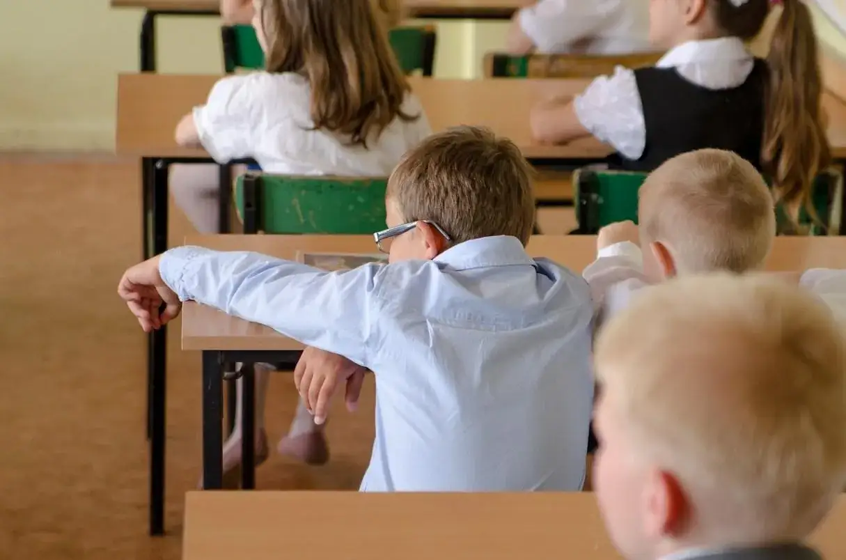 Un bambino iperattivo di 7 anni, con occhiali, si appoggia al banco in classe, annoiato.
