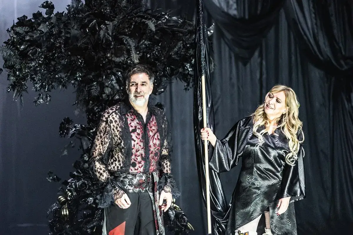 Dwóch doświadczonych aktorów seniorów na scenie teatru. Mężczyzna w ozdobnej koszuli, kobieta trzyma czarną flagę.