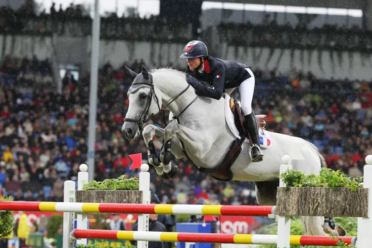 Le cheval Mylord Carthago et sa cavalière sautent un obstacle sous la pluie, devant une foule attentive.