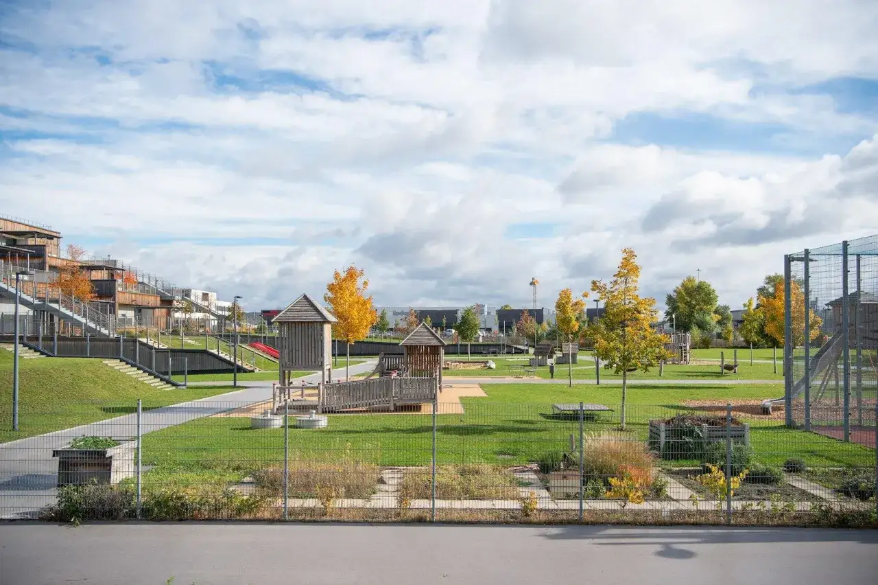 Spielplatz mit Holzspielh&auml;usern, Rutschen und Gr&uuml;nfl&auml;chen. Moderne Geb&auml;ude im Hintergrund. Ein Beispiel f&uuml;r gendergerechte Stadtplanung, die allen zugutekommt.