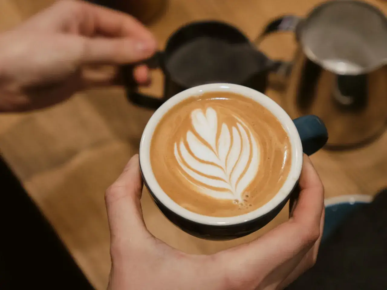 Eine Hand hält eine Tasse mit einem kunstvollen Latte Art Muster, das an ein Blatt erinnert. Daneben stehen Milchkännchen, bereit für den perfekten Cappuccino mit Filterkaffee.