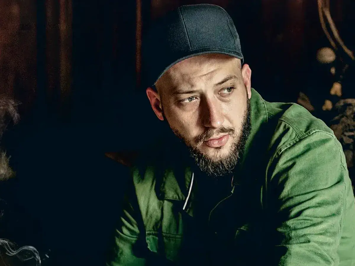 Mężczyzna w czapce i zielonej kurtce, z brodą, wygląda jakby myślał o swojej żonie.