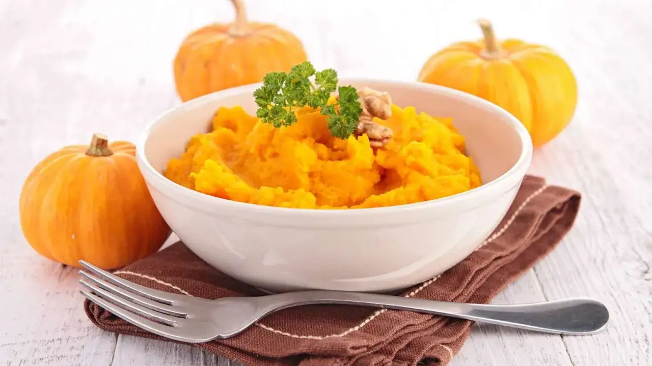 Un tazón de cremoso puré de calabaza, decorado con perejil y nueces, junto a calabazas enteras.