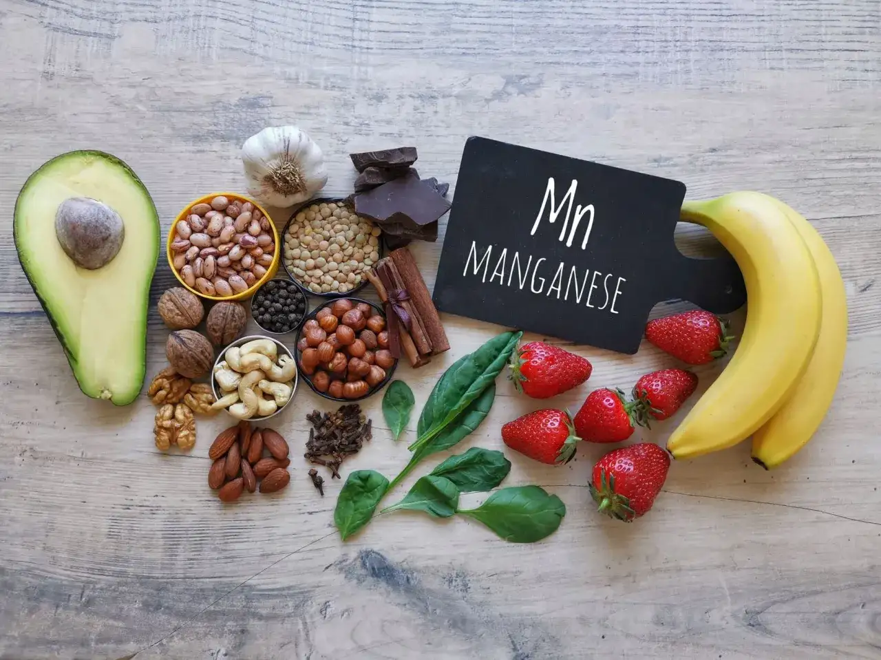 Na drewnianym blacie ułożono produkty bogate w mangan: awokado, fasolę, orzechy, migdały, truskawki, banany, szpinak, czosnek, czekoladę i przyprawy. Tabliczka z napisem 