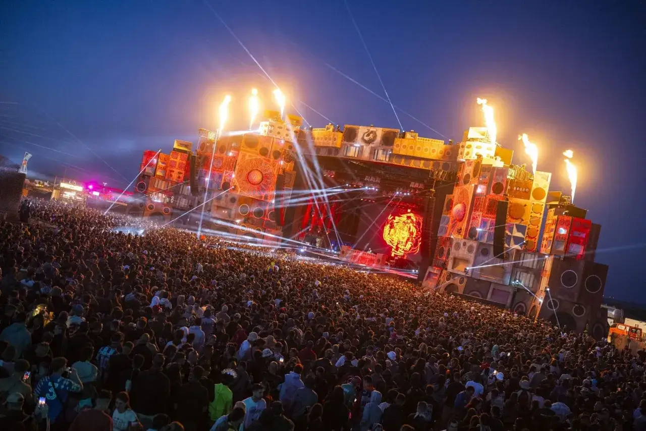 Un'enorme folla balla sotto un palco imponente, illuminato da fiamme e luci laser, un'esperienza immersiva di vari tipi di techno.