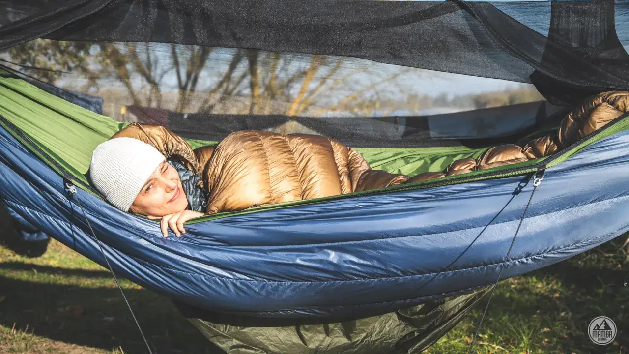 Kobieta w śpiworze i czapce odpoczywa w hamaku, przygotowując się na nocowanie w lesie.