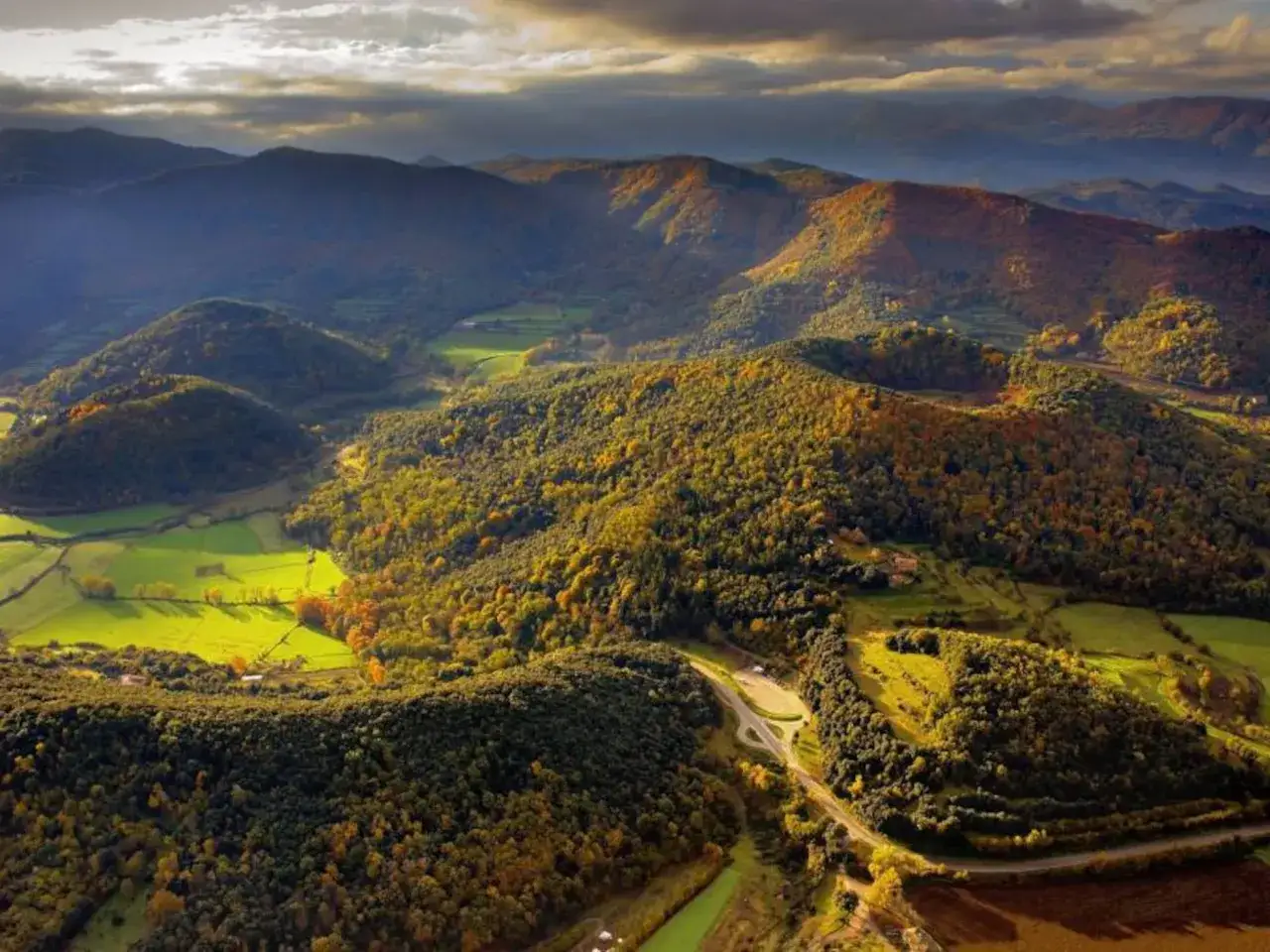 Paisaje otoñal en **La Garrotxa**, con colinas cubiertas de bosques de colores cálidos y campos verdes bajo un cielo nublado.