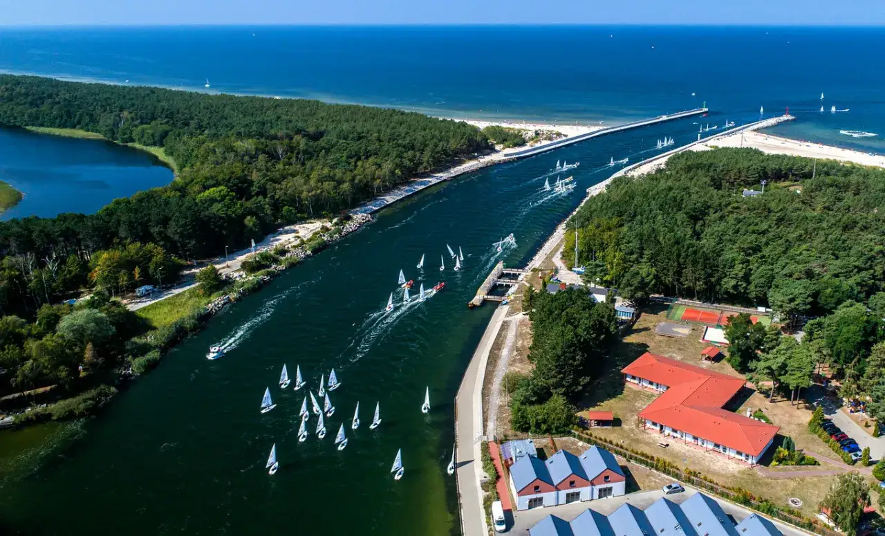 Regaty żaglówek na kanale, otoczonym lasem i wodą. W oddali widać morze i plażę. To jedne z wielu atrakcji Dziwnowa.