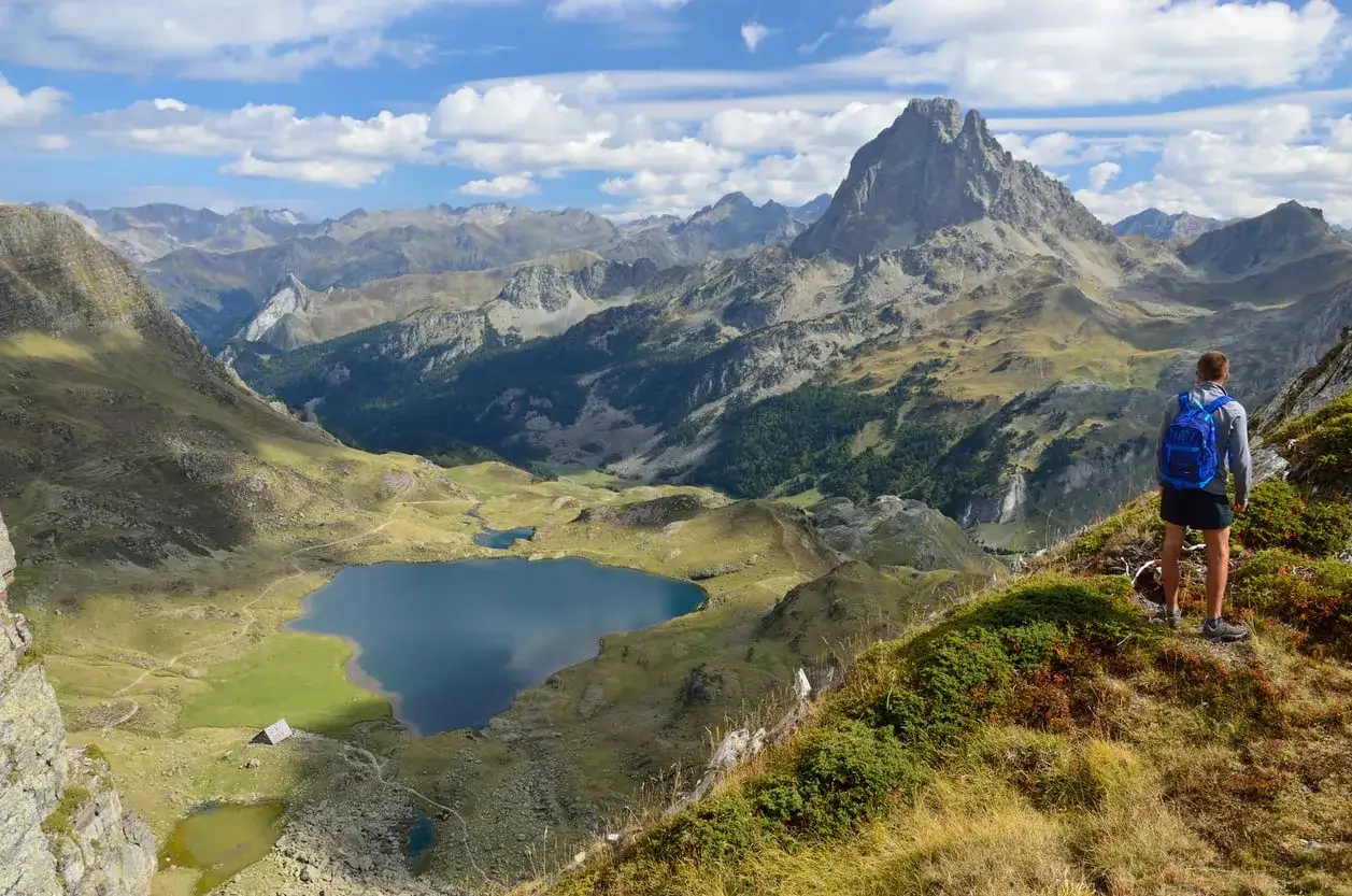 A hiker with a blue backpack surveys a stunning valley with lakes and jagged peaks, a classic view on the Haute Route Pyrenees.