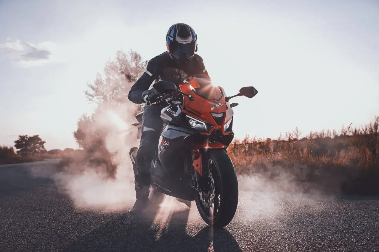 Motocyklista w kasku i kombinezonie na pomarańczowym motocyklu Barton Blade, z kt&oacute;rego unosi się dym. Słońce przebija się przez zadymienie.
