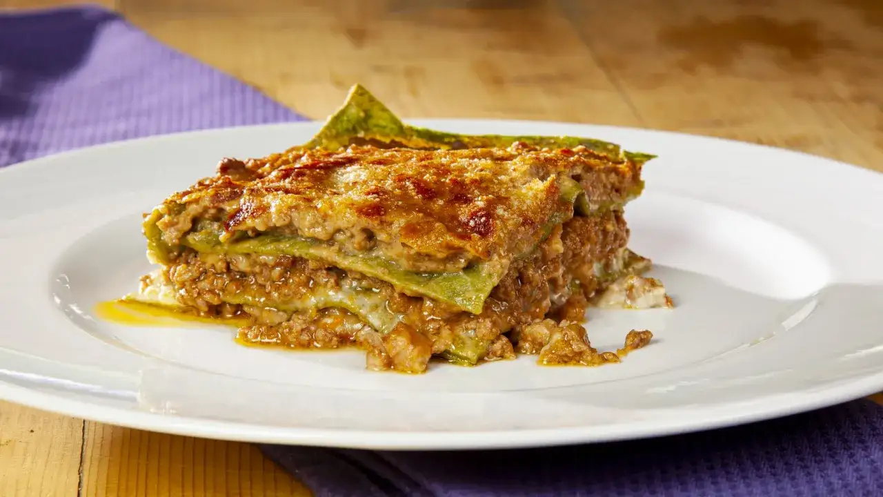 Una fetta di lasagne alla bolognese, con strati di pasta verde, ragù e formaggio fuso, servita su un piatto bianco.