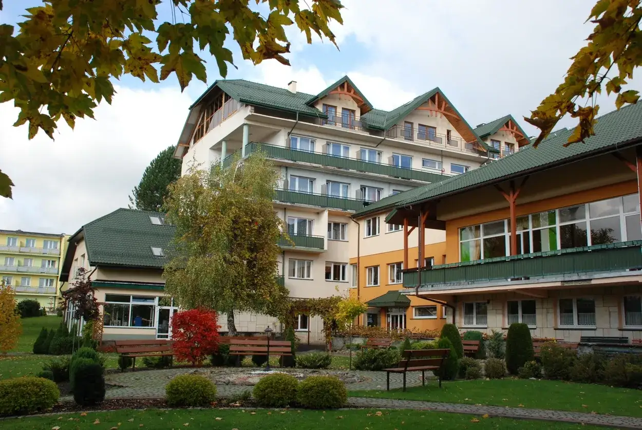 Sanatorium Plon Polańczyk w jesiennej odsłonie. Budynki z zielonymi dachami, otoczone zadbanym ogrodem z ławkami i drzewami.