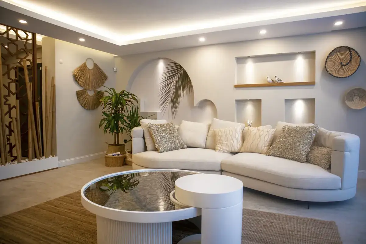 Sofá curvo beige con cojines decorativos, mesa de centro redonda, alfombras de yute y elementos decorativos de bambú y paja. La iluminación indirecta crea un ambiente acogedor, ideal para combinar lámparas salón comedor.